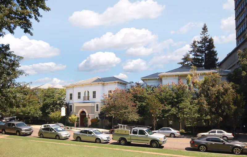 View of a street with parked cars in front of a two-story building surrounded by trees under a partly cloudy sky.