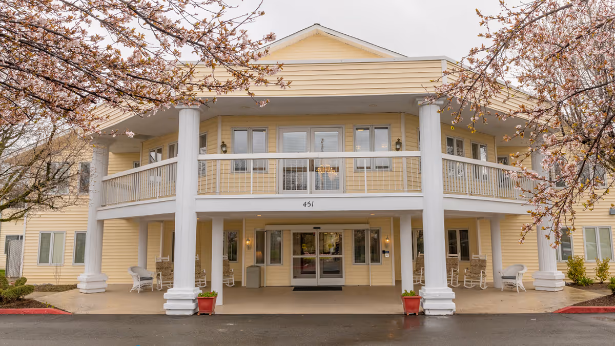 Front exterior view of a two-story senior living facility building with yellow siding, white columns, and a balcony. There are rocking chairs on the covered porch area and blossoming trees framing the entrance.