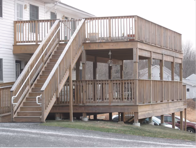 Two-level wooden deck attached to a white house with stairs leading from the ground to the upper deck. The deck has wooden railings and is elevated on wooden posts. There is a grassy area and a paved driveway in the foreground, with some houses and trees visible in the background.