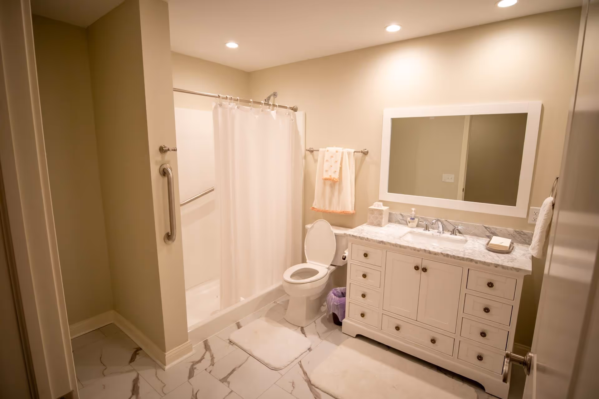 A clean and well-lit bathroom featuring a white toilet with the lid open, a white vanity with a marble countertop and a large mirror above it, a shower with a white curtain, beige walls, and marble-patterned floor tiles. Towels hang on racks near the shower and vanity.