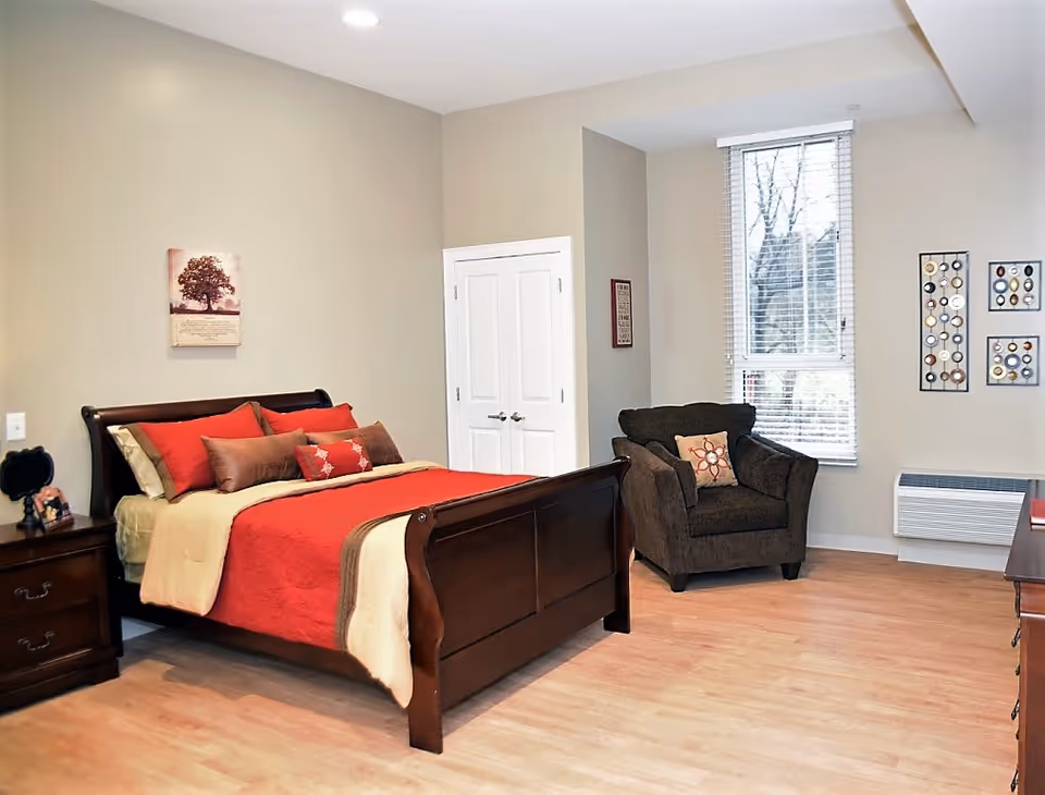 Well-lit bedroom with a wooden bed dressed in red and beige bedding, a nightstand, an upholstered armchair, and a window.
