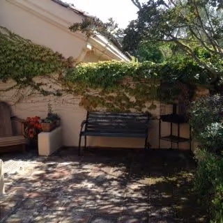 Outdoor patio area with a black metal bench, a wooden chair, and a small table. The wall behind is covered with green ivy, and there are trees providing shade. The ground is paved with stone tiles.