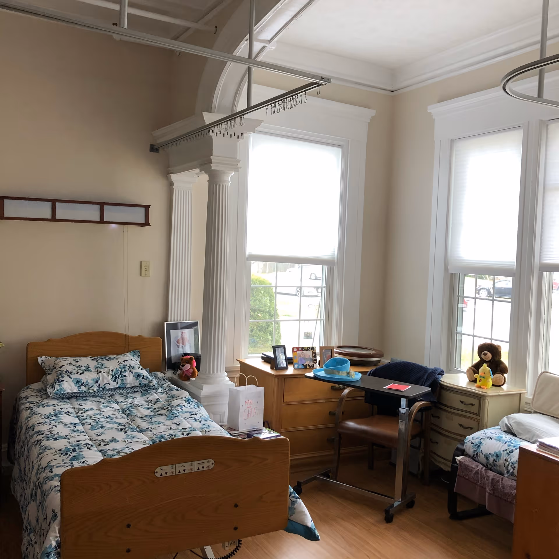 A bright senior-care bedroom with two twin beds, wooden furniture, large windows with blinds, decorative columns, and personal items like stuffed animals and photos.