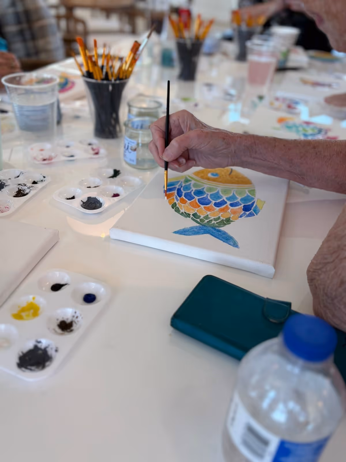 An elderly person painting a colorful fish on a small canvas at a table with paint palettes, brushes in containers, and a water bottle nearby.