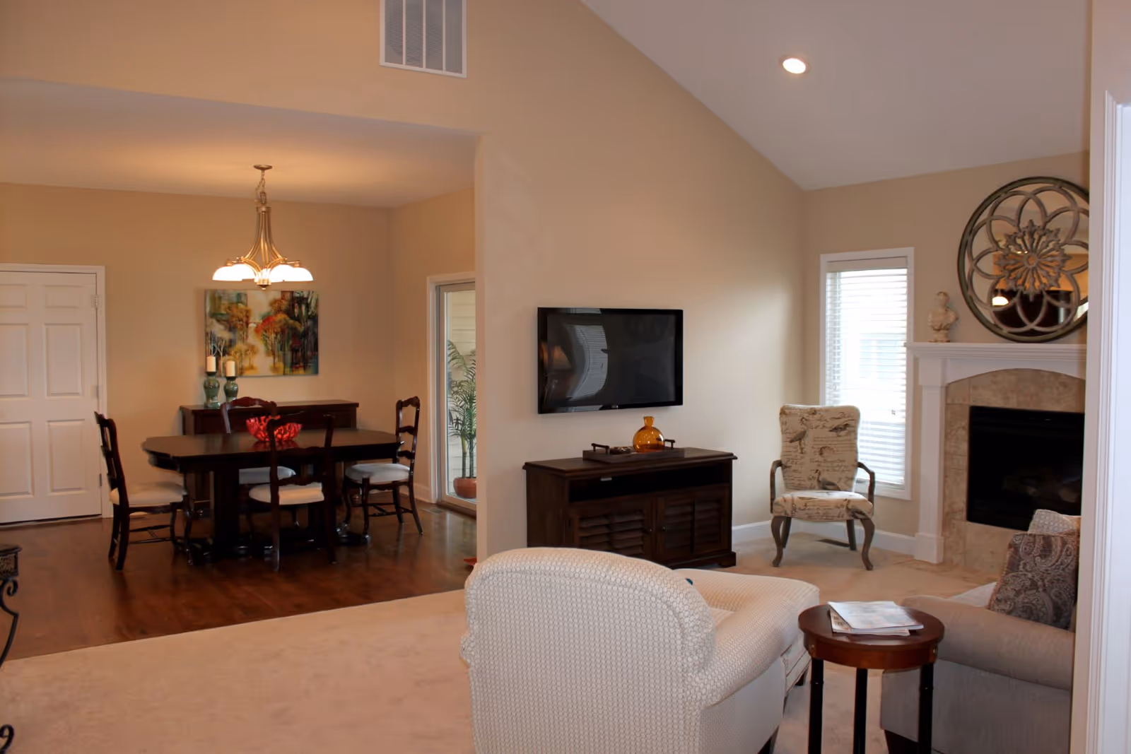 Interior view of a senior living facility showing a living room and dining area. The living room has a beige armchair, a patterned chair, a small round wooden table with magazines, a mounted flat-screen TV above a wooden cabinet, and a fireplace with a decorative round mirror and a small statue on the mantel. The dining area features a dark wooden dining table with six chairs, a sideboard with decorative items, a colorful painting on the wall, and a chandelier overhead. The walls are painted beige, and the floor is a combination of carpet and hardwood.