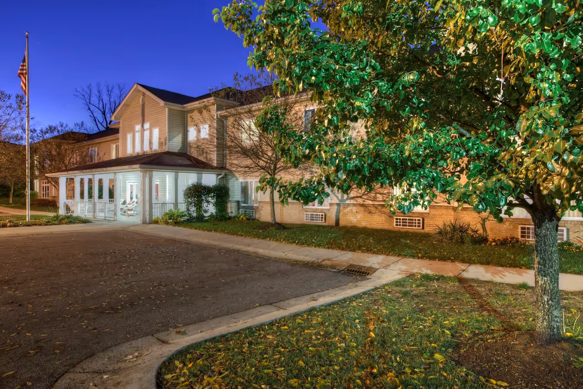 Exterior view of a two-story senior living facility building at dusk with a well-lit entrance, an American flag on a flagpole, a large tree with green leaves, and a paved driveway and sidewalk in front.