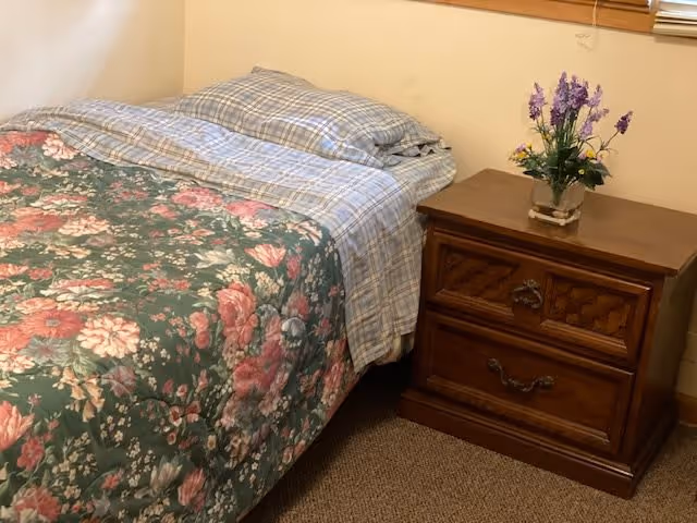 A bedroom corner featuring a bed with a floral patterned comforter and plaid sheets, next to a wooden nightstand with two drawers and a small vase of purple flowers on top.