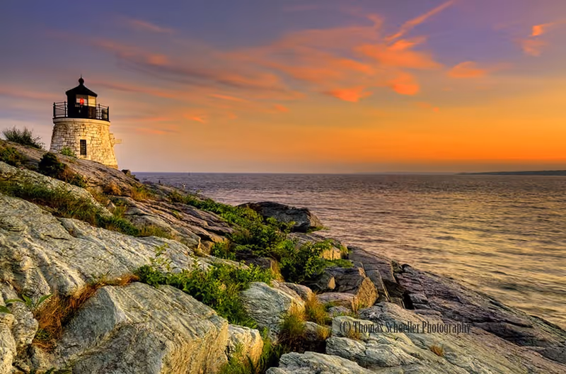 A stone lighthouse on a rocky coastline overlooking the ocean at sunset with an orange and purple sky.