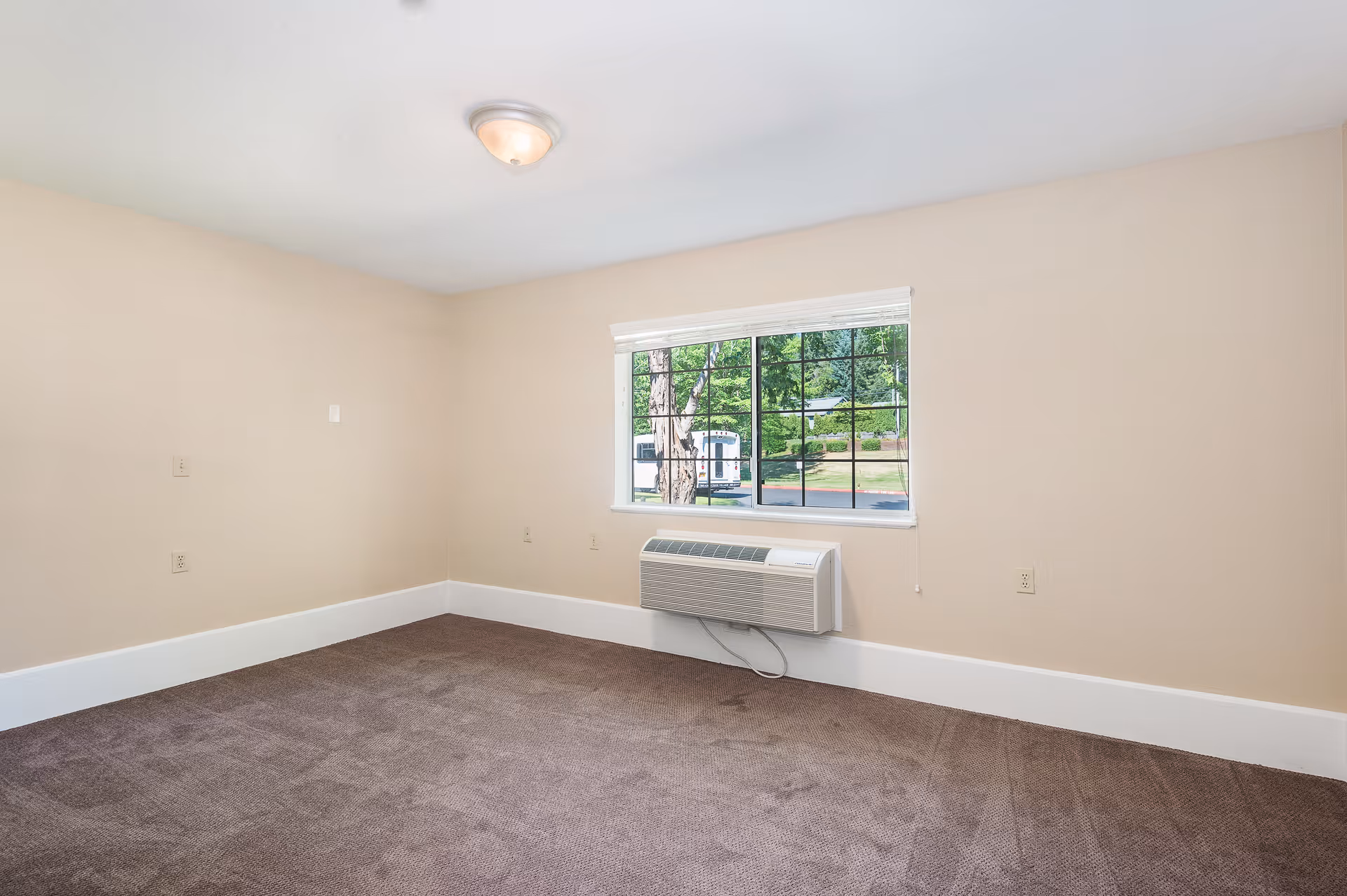 Empty room with beige walls, brown carpet, a window with a view of trees and a bus outside, and a wall-mounted air conditioning unit below the window.