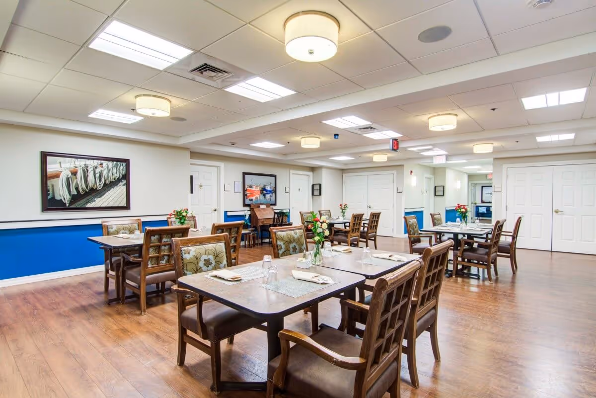 A bright and spacious dining room with several wooden tables and chairs arranged neatly. Each table is set with placemats, napkins, and glasses, and has a small vase with flowers. The room has wood flooring, white walls with a blue accent stripe, and ceiling lights. Nautical-themed artwork is hung on the walls.