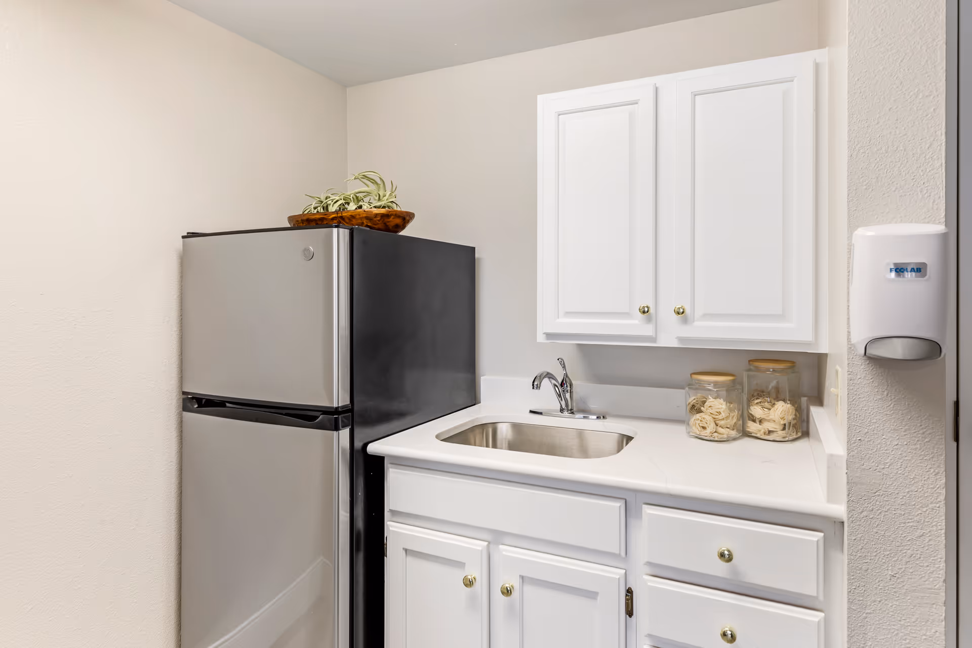 A small kitchen area with a stainless steel refrigerator, white cabinets with gold knobs, a countertop with a built-in sink and faucet, two glass jars with pasta, and a wall-mounted Ecolab soap dispenser.