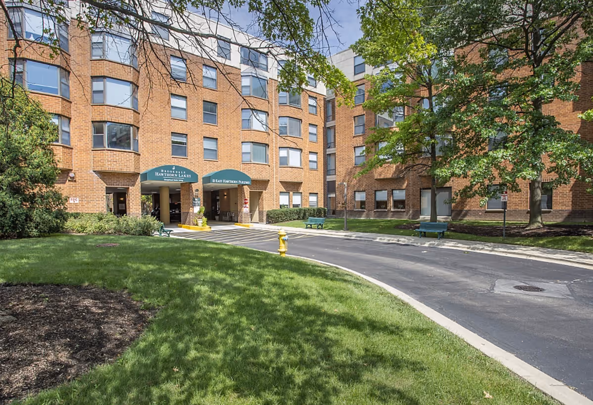 Exterior view of a multi-story brick building with green awnings over the entrances labeled 'Hawthorn Lakes'. The building is surrounded by green grass, trees, and a paved driveway with benches along the sidewalk.