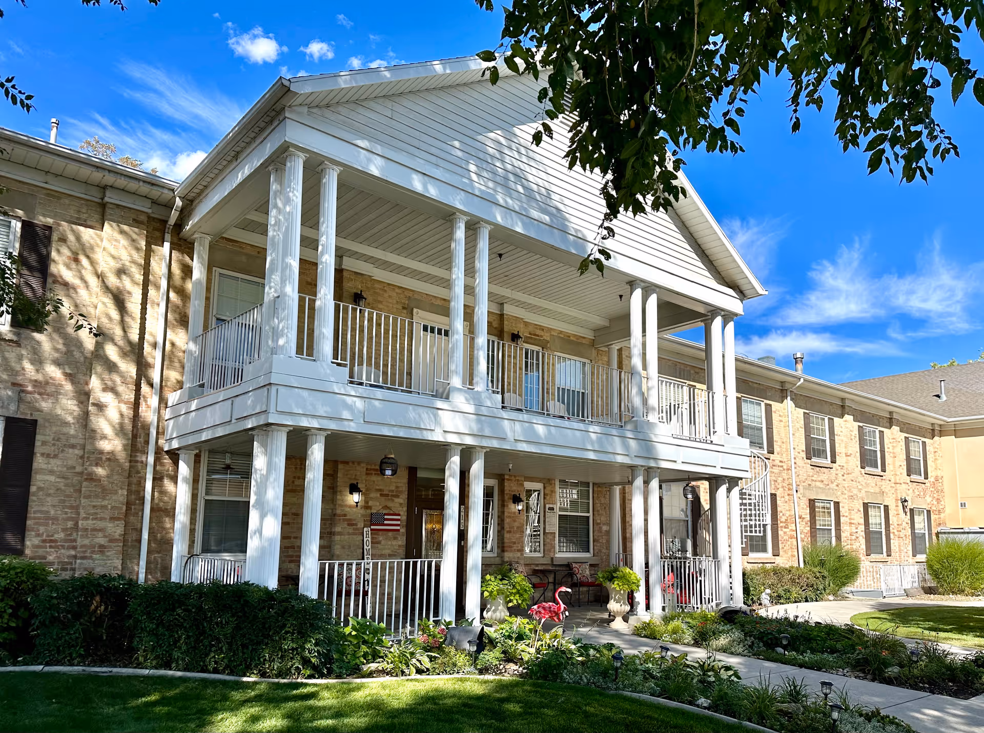 Front exterior of Abbington Manor showing a two-story brick building with white columns, porches, and landscaped lawn under a blue sky.