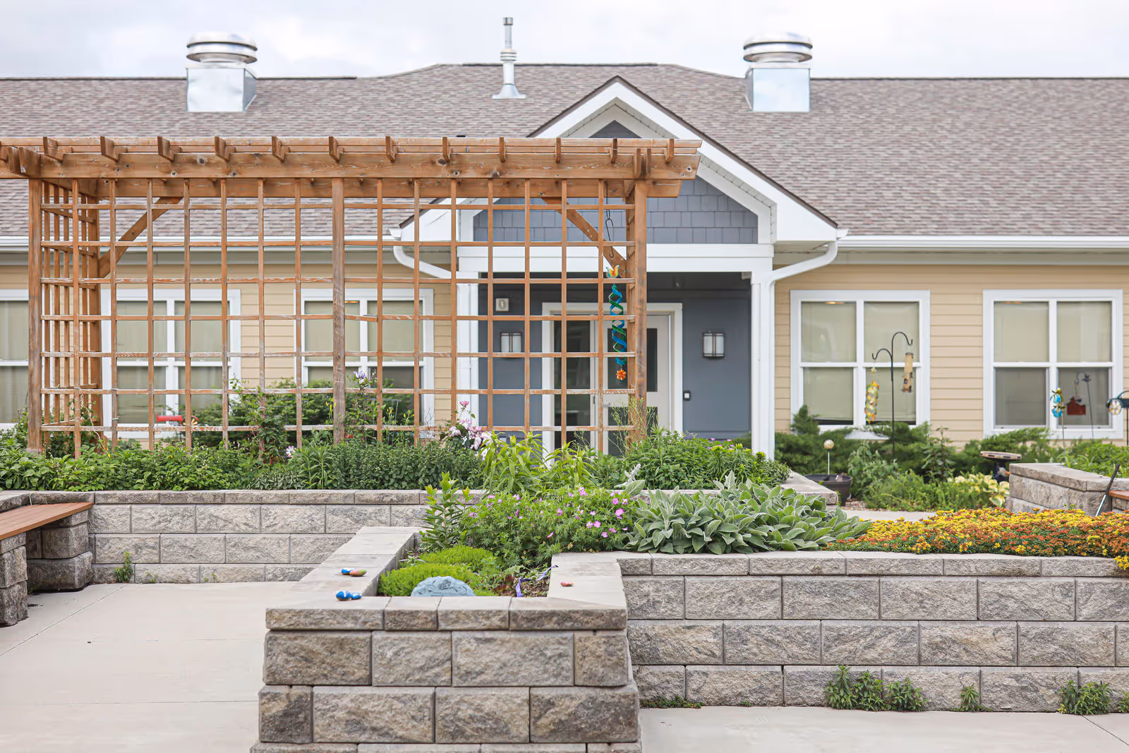 Landscaped courtyard with raised stone planters, a wooden pergola, and the entrance to a single-story senior living building.