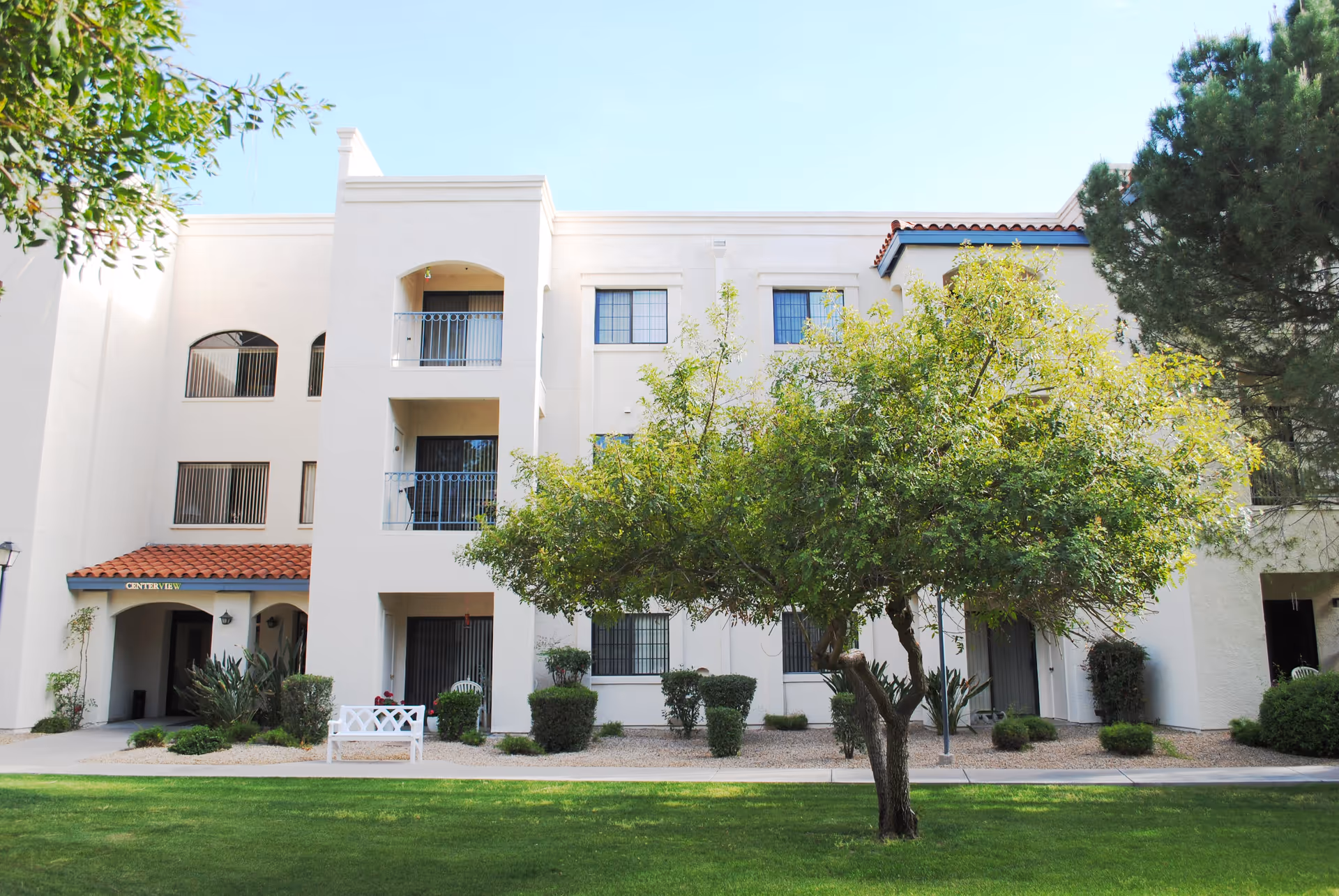 Exterior view of a three-story senior living facility building with white walls and red tile roof accents. There are balconies and windows visible, along with a green lawn, a tree, shrubs, and a white bench in the foreground under a clear blue sky.