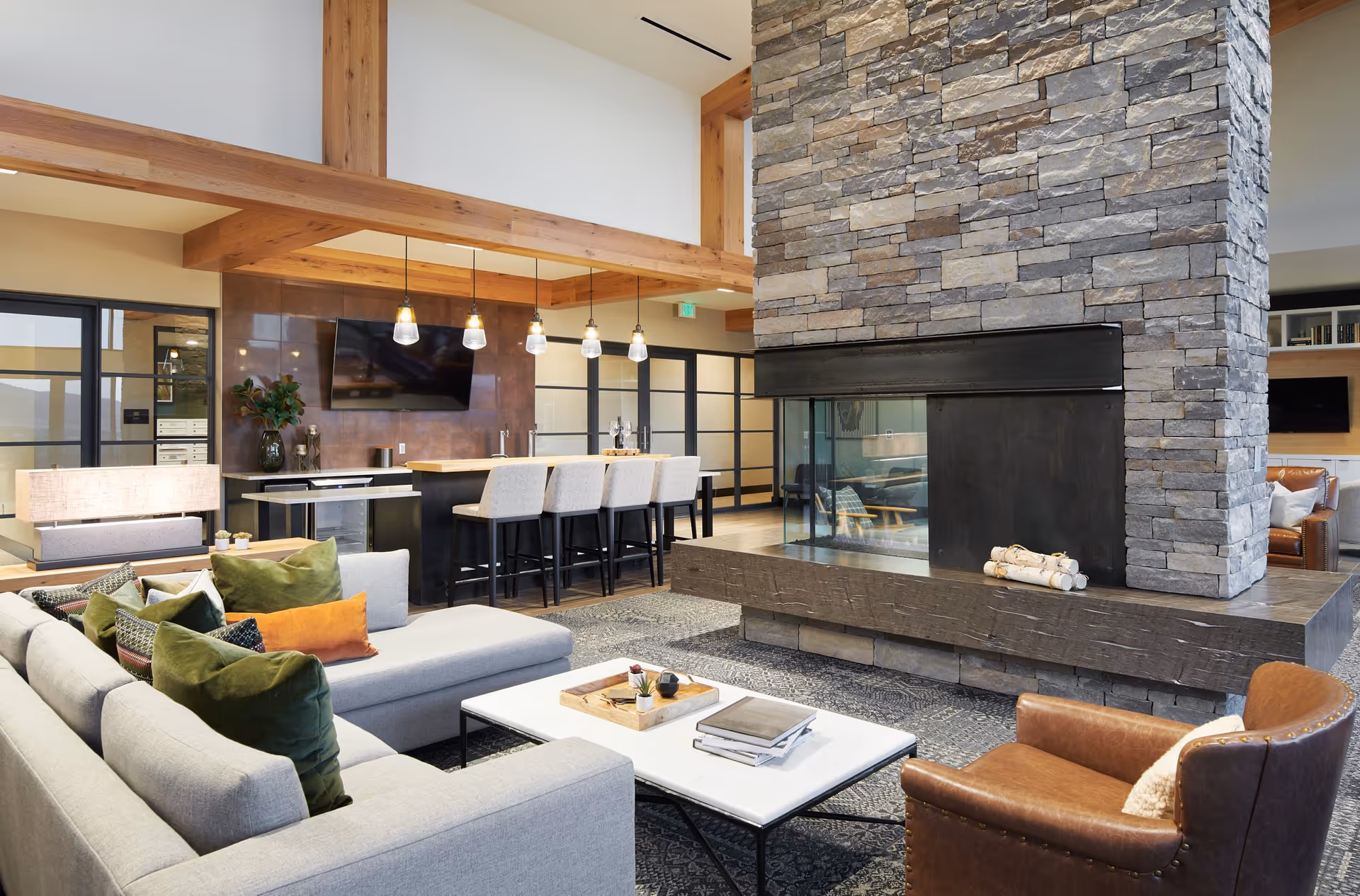 A modern and cozy living room area with a large stone fireplace in the center. The room features a light gray sectional sofa with green and orange pillows, a white coffee table with books and a tray, a brown leather armchair, and a bar area with four high chairs and pendant lights hanging above. There is a flat-screen TV mounted on the wall behind the bar, and large windows and glass doors in the background.