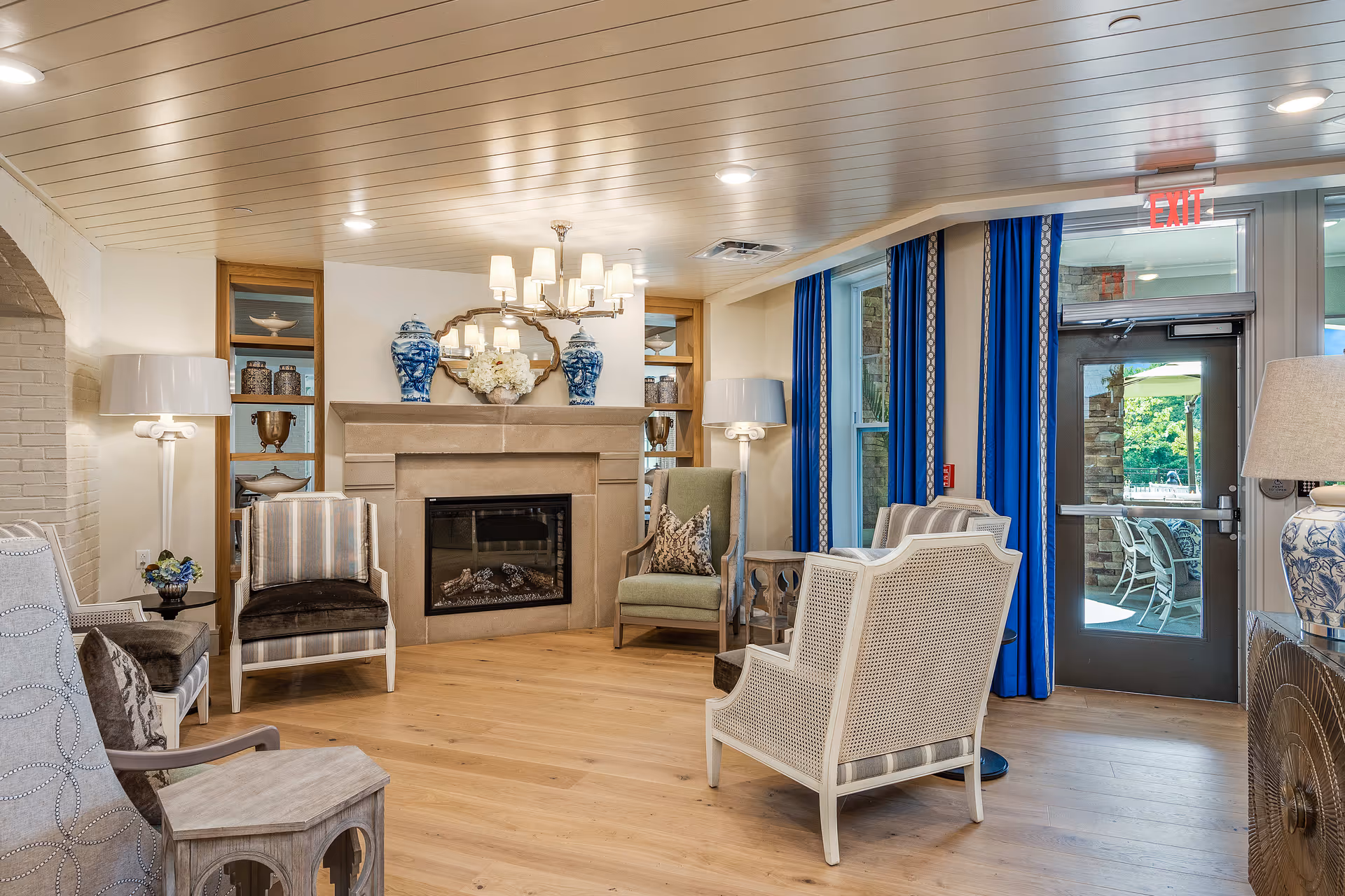 A cozy living room area with a stone fireplace decorated with blue and white vases and a floral arrangement. The room features several upholstered chairs arranged around the fireplace, wooden flooring, blue curtains on the windows, and a glass door leading outside. The ceiling has recessed lighting and a chandelier above the fireplace.