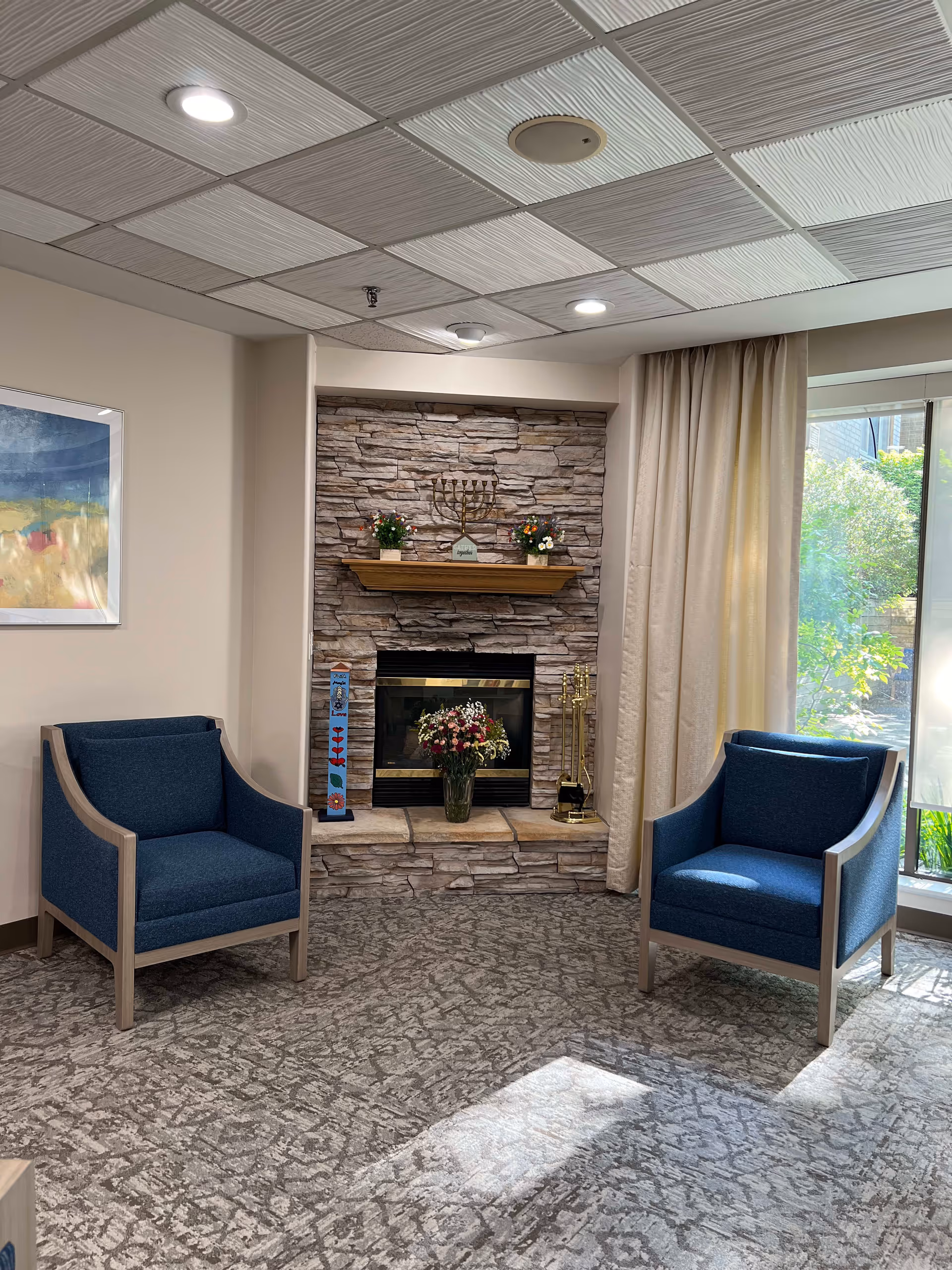 A cozy sitting area with two blue armchairs facing a stone fireplace. The fireplace mantel holds a menorah and two flower arrangements. There is a vase with flowers on the hearth. A large window with beige curtains lets in natural light, and a framed abstract painting hangs on the wall to the left.