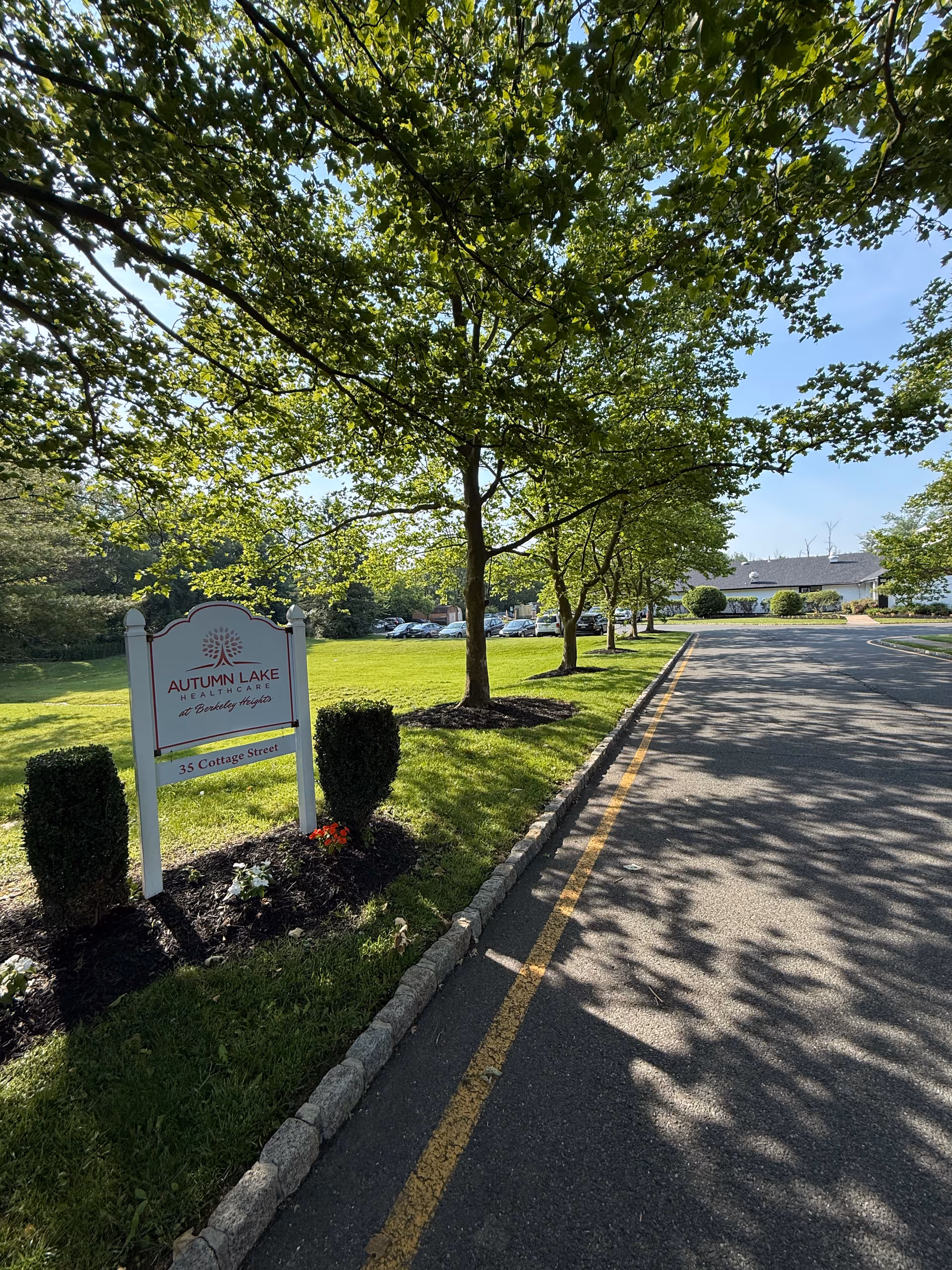 An entrance sign for Autumn Lake Healthcare stands on a grassy lawn beside a tree-lined driveway leading to the facility building.