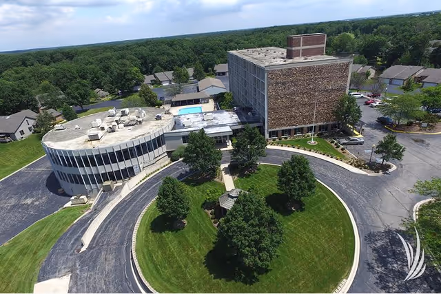 Aerial view of Village Woods senior living facility showing a circular driveway with green lawn and trees in the center, a multi-story building with a brick facade, a curved glass-fronted building, parking areas, and a swimming pool in the background surrounded by trees and residential houses.