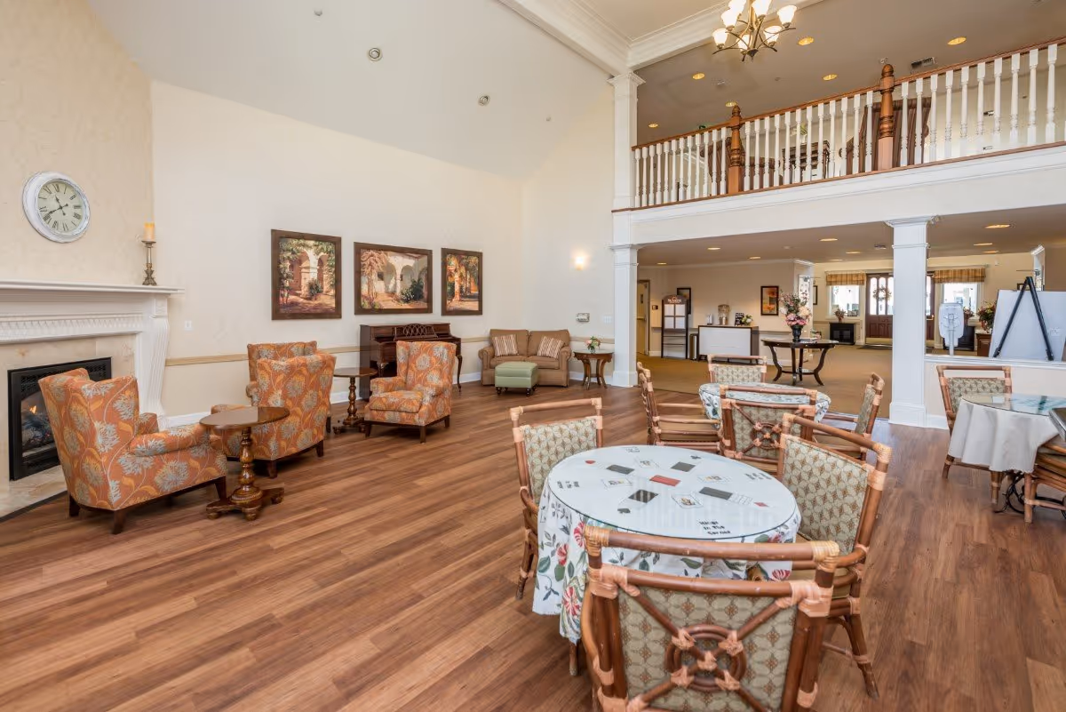 A spacious senior living common area with wooden flooring, several round tables with chairs, and a seating area with floral upholstered armchairs and a small sofa. The room features a fireplace with a clock above it, three framed paintings on the wall, and a piano. There is a balcony railing on the upper level and a reception desk visible in the background.