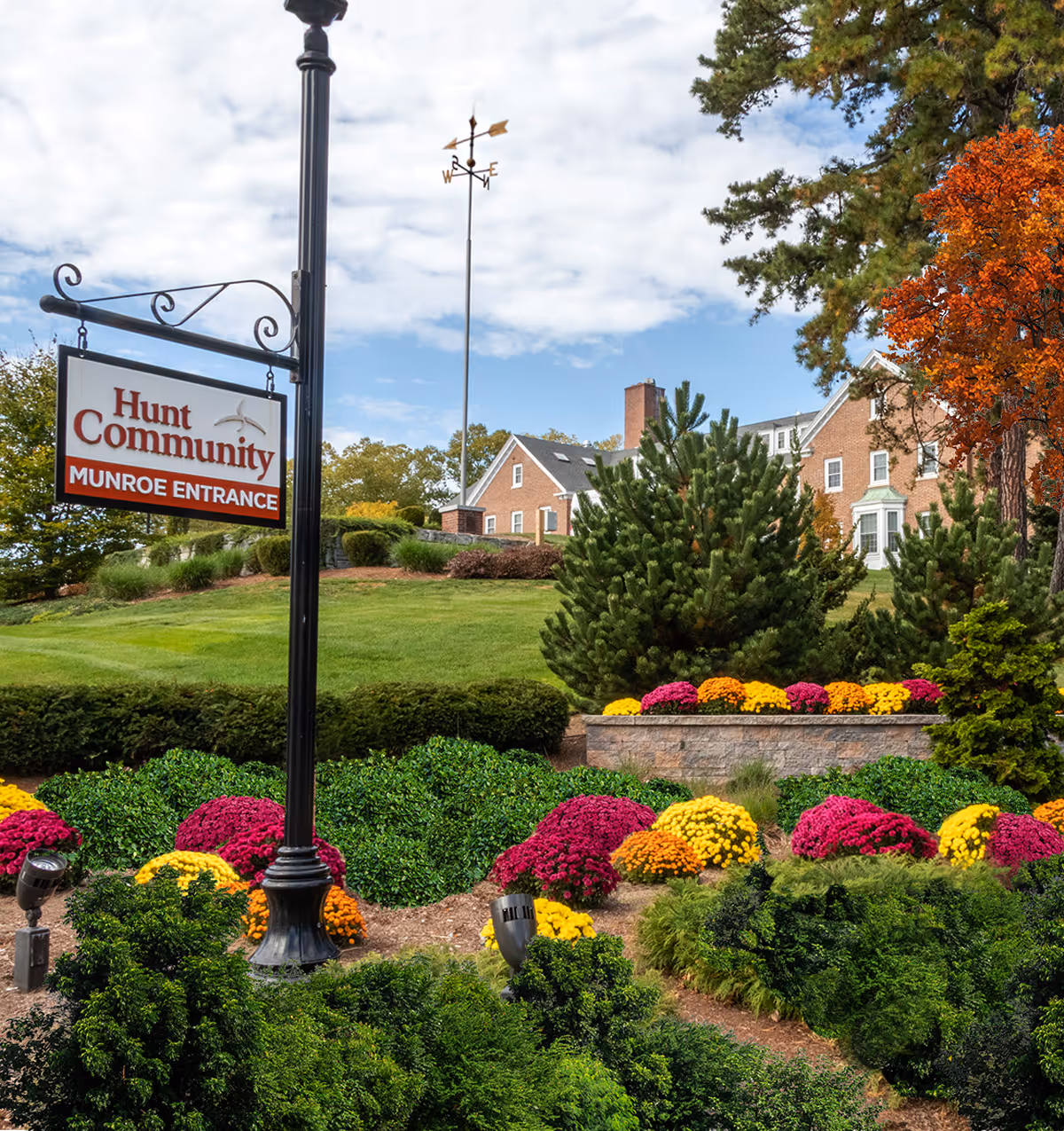 Outdoor view of Hunt Community Munroe Entrance with a landscaped garden featuring colorful flowers and green shrubs. A brick building is visible in the background under a partly cloudy sky.