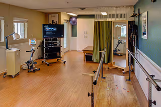 Empty rehabilitation gym with parallel bars, exercise equipment, TV, and a privacy curtain.