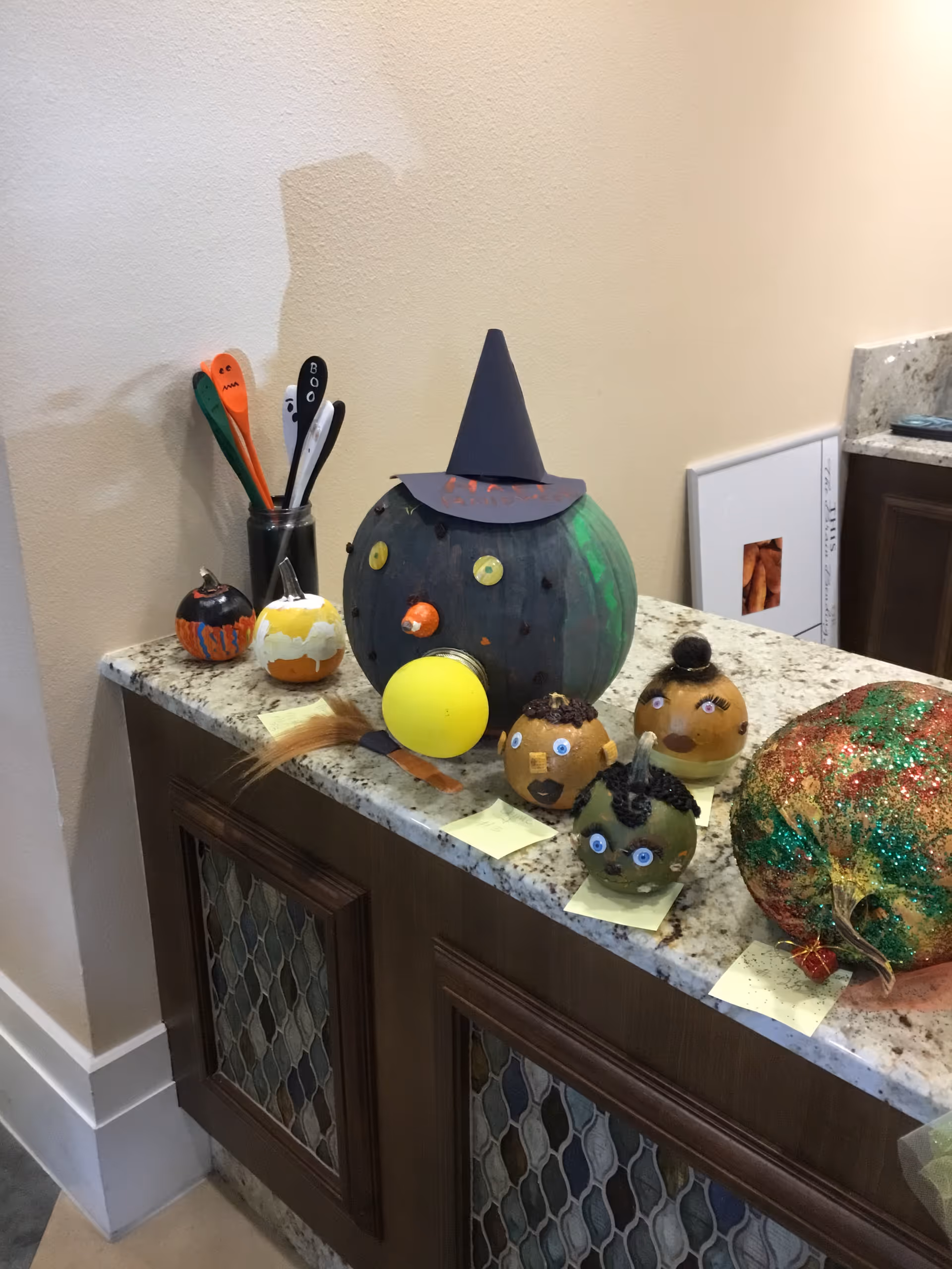 A granite countertop with several decorated pumpkins and gourds arranged on it. The largest pumpkin is painted dark green with a witch hat and has a yellow ball attached to the front. Smaller pumpkins and gourds are decorated with faces and various materials. There are also some colorful spoons in a jar on the left side of the counter. The countertop is part of a wooden cabinet with decorative glass panels.