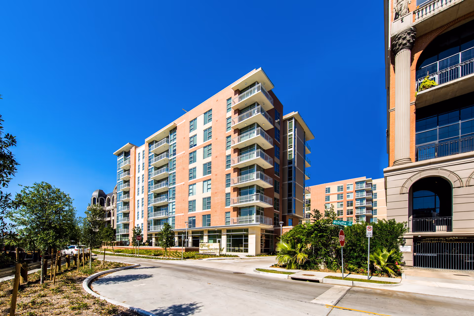 Exterior view of a modern multi-story residential building with balconies under a clear blue sky. The building is surrounded by trees and a curved driveway leading to the entrance. A street sign and stop sign are visible near the entrance.