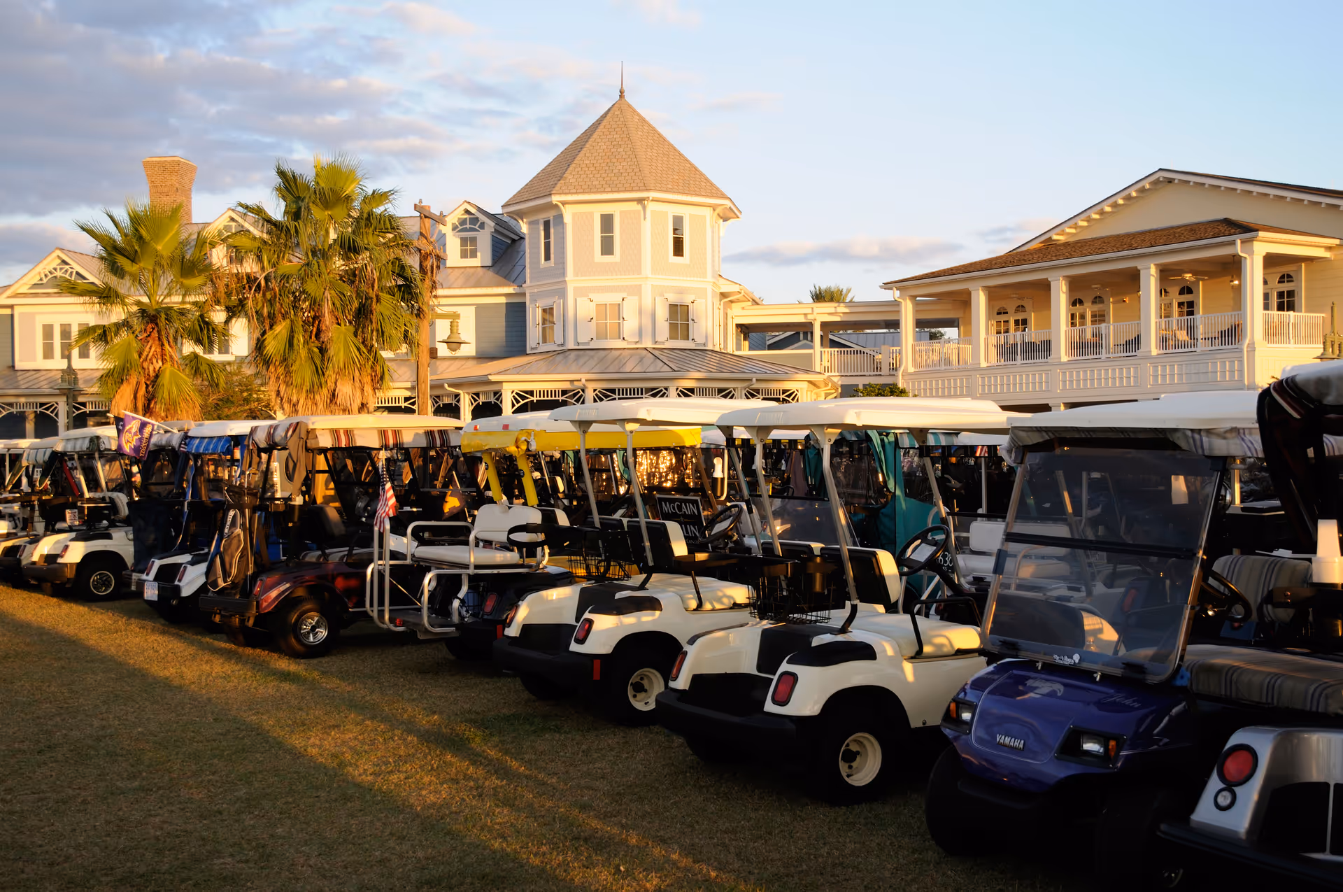 A row of parked golf carts on grass in front of a large, elegant building with a turret and wraparound porch, illuminated by warm sunlight under a partly cloudy sky.