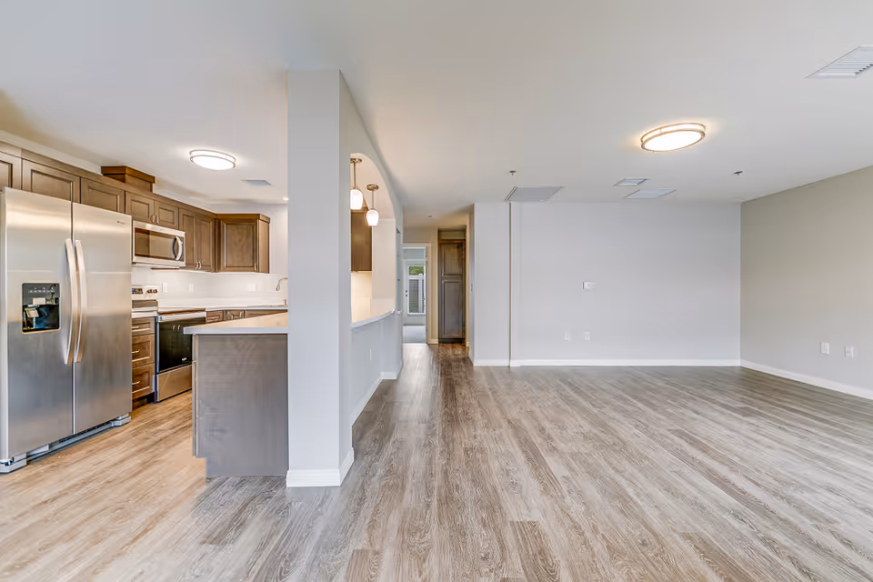 Open-plan interior showing a modern kitchen with stainless steel appliances opening into a spacious empty living area with wood floors.