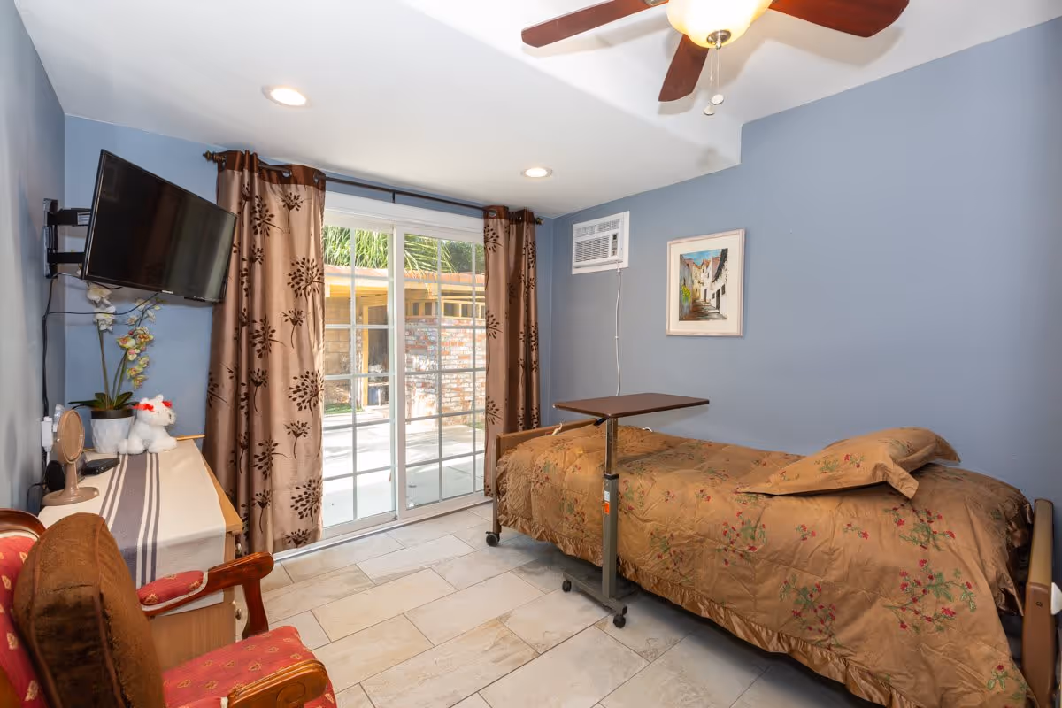 A small bedroom with light blue walls featuring a single bed with a brown floral bedspread and pillow. There is a brown adjustable overbed table on wheels next to the bed. A wooden chair with red upholstery and a brown cushion is placed near a wooden dresser covered with a striped cloth, holding a small mirror, a potted orchid, and a white stuffed animal. A flat-screen TV is mounted on the wall above the dresser. The room has a ceiling fan with a light, recessed ceiling lights, and a window air conditioning unit. Sliding glass doors with brown patterned curtains open to an outdoor area with a brick wall and greenery.