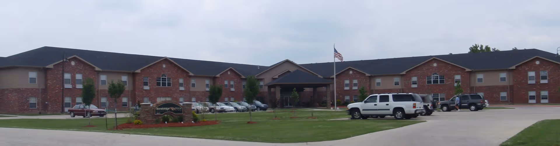 Wide exterior view of a two-story brick senior living facility named Stonebridge Place with a parking lot in front, several cars parked, a flagpole with an American flag, and a well-maintained lawn with small trees and landscaping.