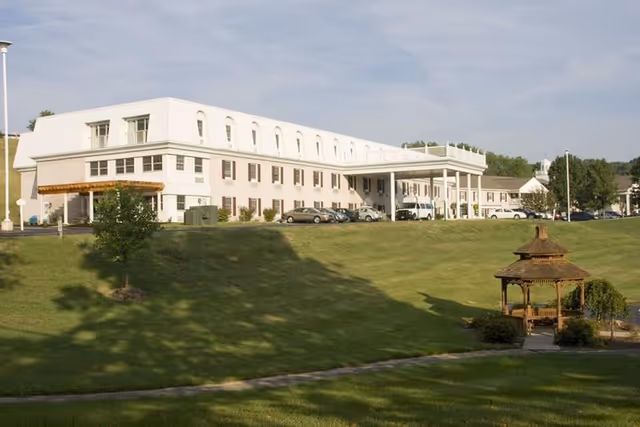 Front view of a multi-story white senior living building with a covered entrance, parked cars, a wide grassy lawn, and a wooden gazebo.