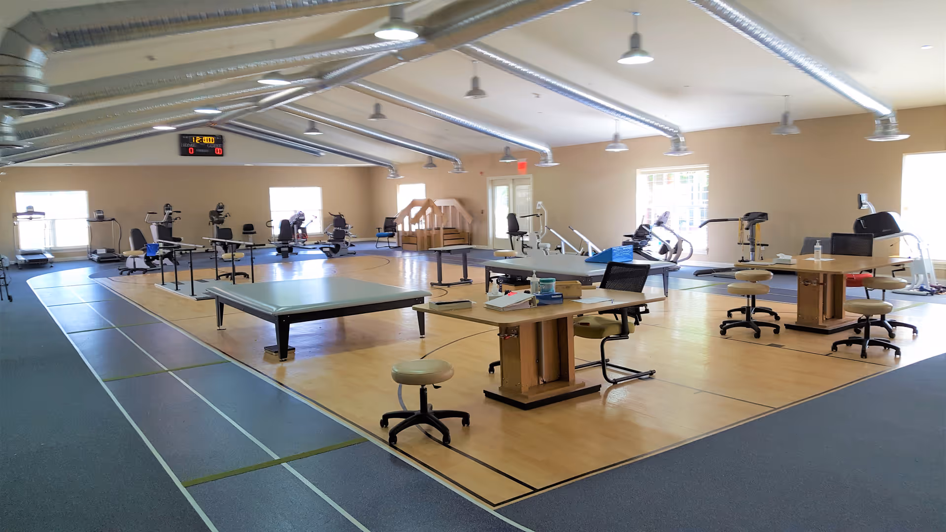 A spacious rehabilitation or physical therapy room with exercise equipment including treadmills, stationary bikes, parallel bars, therapy tables, and stools. The room has a wooden floor area surrounded by a blue track, large windows letting in natural light, and exposed ductwork on the ceiling.