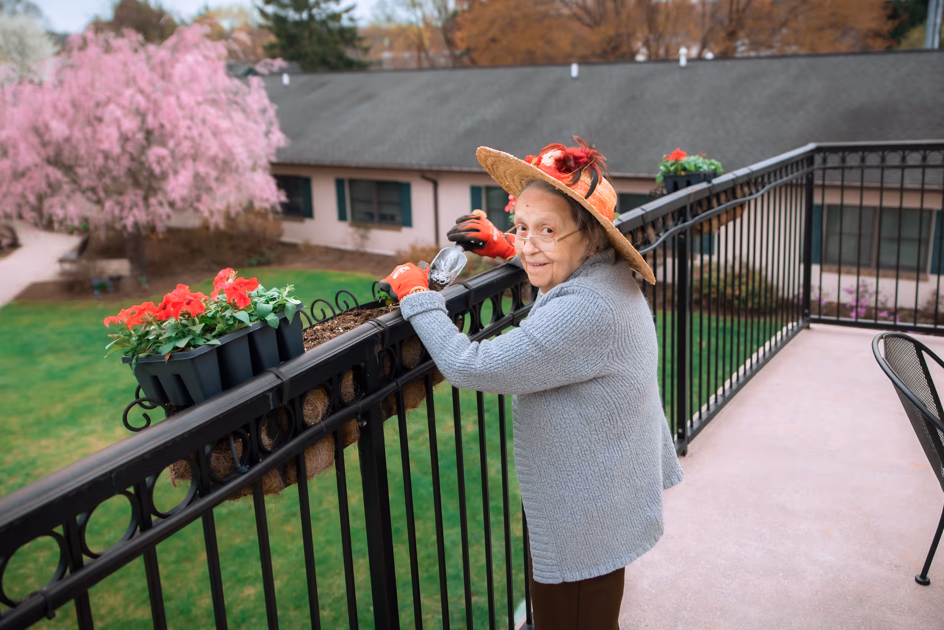 An elderly woman wearing a gray sweater, red gloves, and a decorative straw hat is gardening on a balcony. She is tending to red flowers planted in a railing planter, with a green lawn and a building with green shutters visible in the background.
