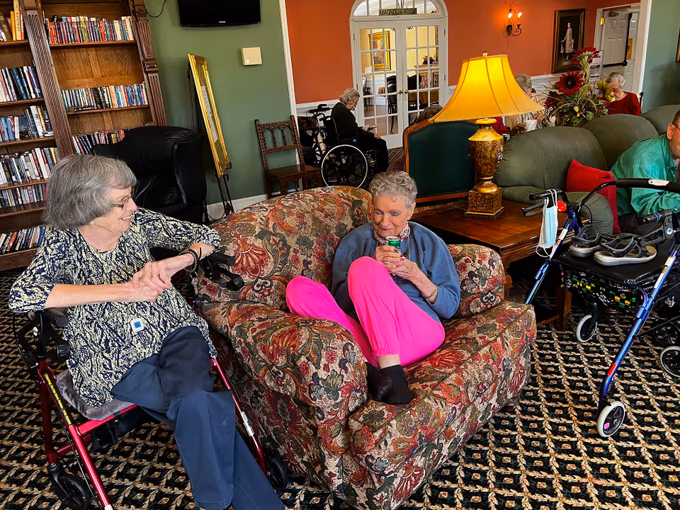 A cozy senior living facility common area with elderly residents sitting and relaxing. One woman in a patterned top and dark pants sits on a walker chair, smiling and talking to another woman in bright pink pants and a blue top who is seated on a floral-patterned armchair holding a drink can. In the background, other residents are seated on couches and chairs near a wooden bookshelf filled with books, a table lamp, and a floral arrangement. The room has warm-colored walls and carpeted flooring with a patterned design.