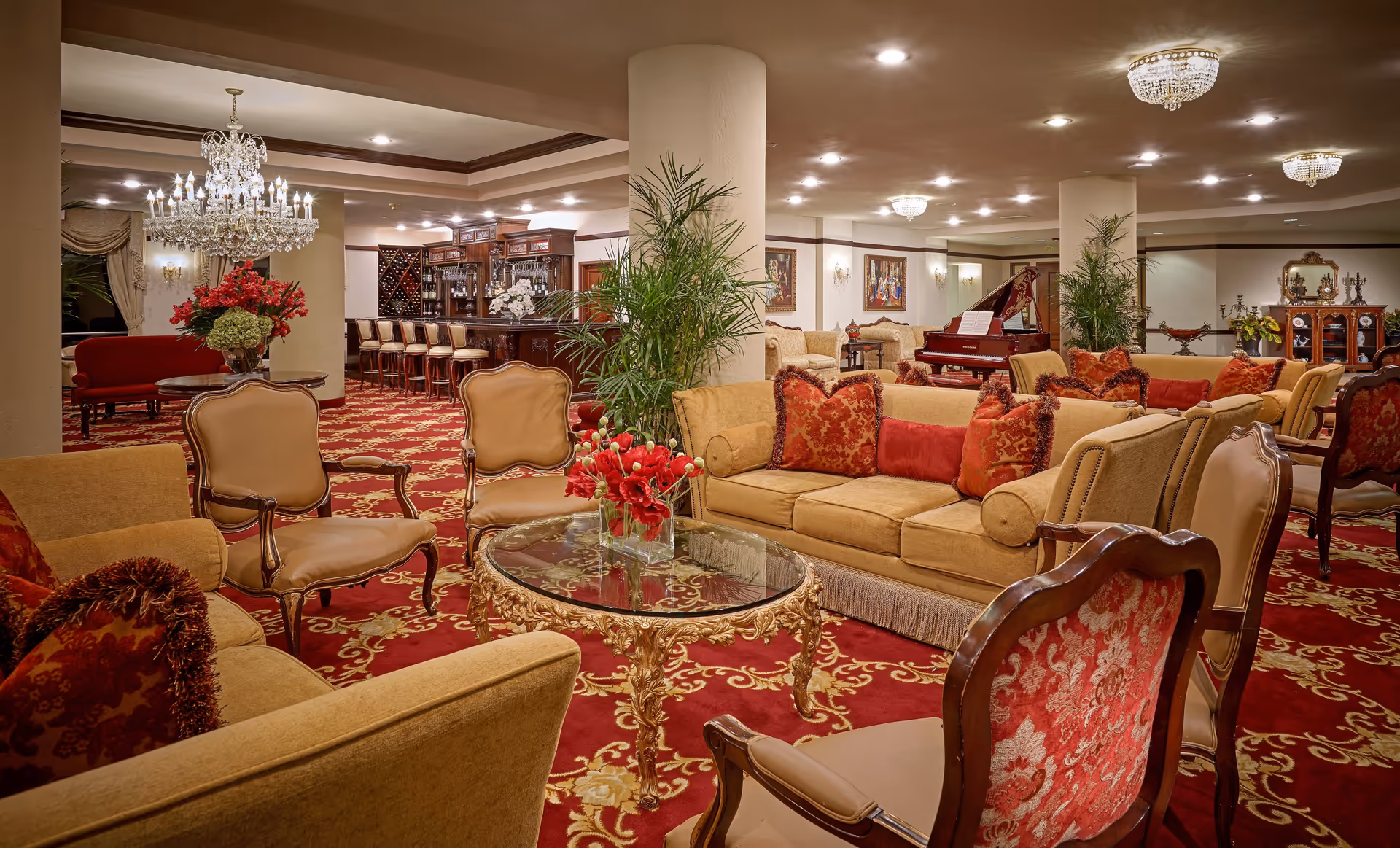 Luxurious living room area with ornate furniture including beige sofas and armchairs with red and gold cushions, a glass-top coffee table with gold detailing, red and gold patterned carpet, large green plants, a grand piano in the background, and a bar area with high chairs under a crystal chandelier.