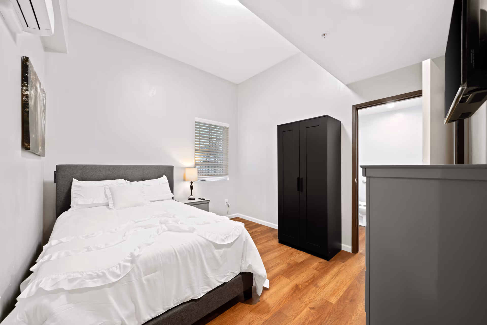 Bright minimalist bedroom with a made bed, bedside table and lamp, black wardrobe, dresser with wall-mounted TV, and hardwood floors.