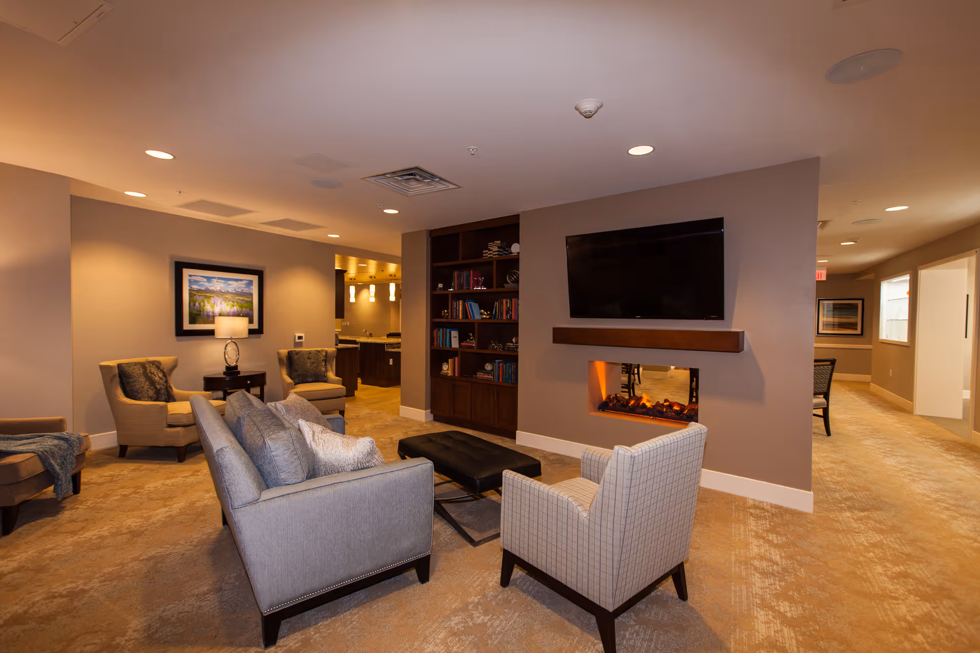 A cozy living room area in a senior living facility with a gray sofa, two armchairs, a black ottoman, and a built-in bookshelf. A flat-screen TV is mounted above a modern electric fireplace. The room is warmly lit with recessed ceiling lights and a table lamp, and there is a hallway visible in the background.