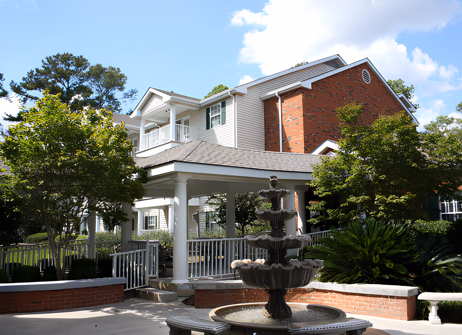 Exterior view of a senior living facility named Langdale Place featuring a multi-story building with white siding and red brick accents, surrounded by green trees and shrubs. In the foreground, there is a covered porch area with white columns and a multi-tiered water fountain. The sky is blue with some clouds.
