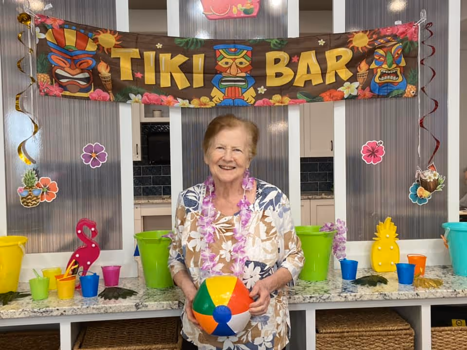 An elderly woman wearing a floral shirt and a purple lei stands smiling in front of a counter decorated with colorful cups, buckets, and tropical-themed items. Above her is a banner that reads 'TIKI BAR' with tiki mask illustrations. The setting appears festive and tropical-themed.