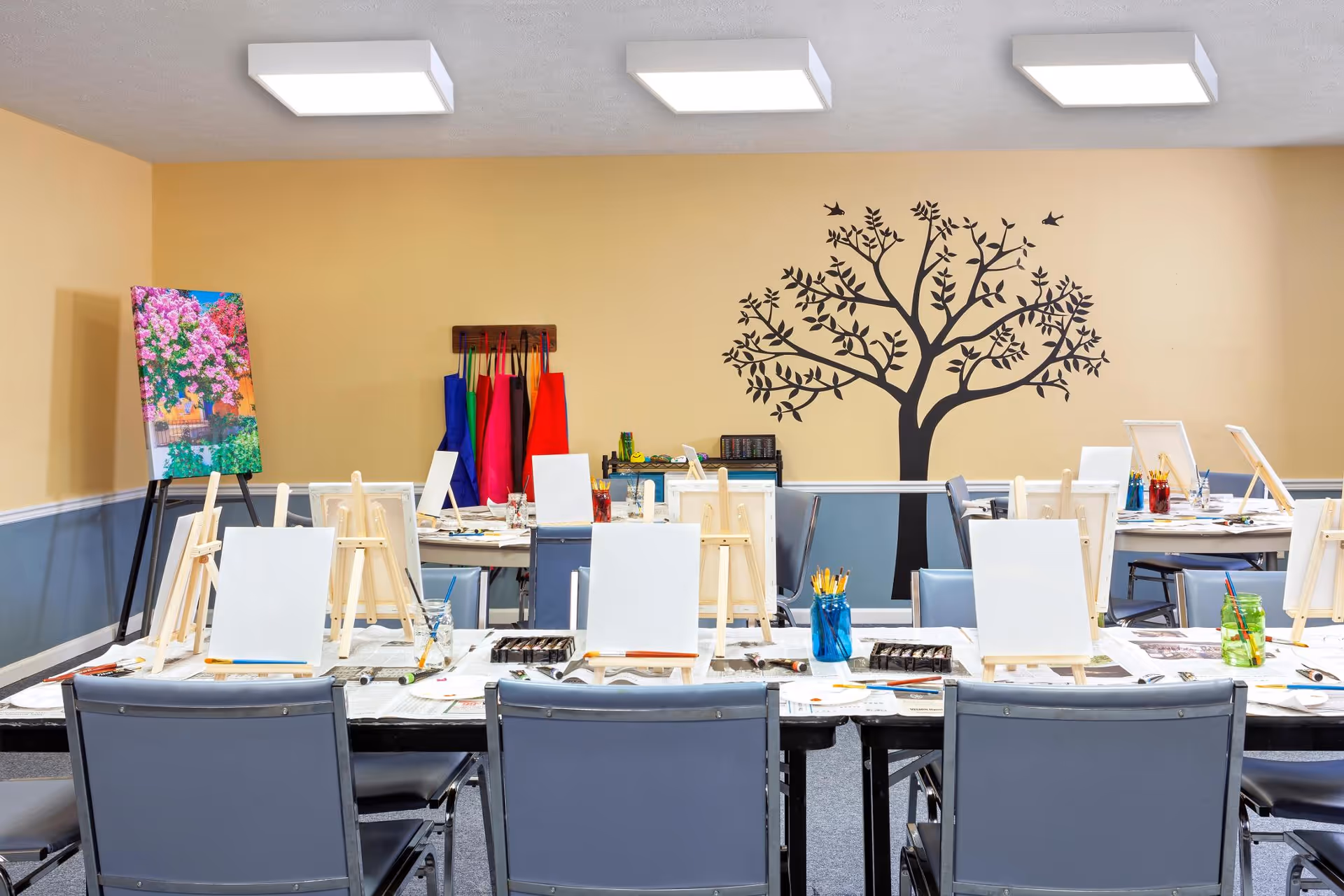 A bright art room with multiple tables arranged for painting activities. Each table has easels with blank canvases, paintbrushes, jars with water, and painting supplies. The walls are painted beige and blue with a large black tree decal and birds on one wall. Colorful aprons hang on hooks, and a finished painting of pink flowers on an easel is visible in the corner.