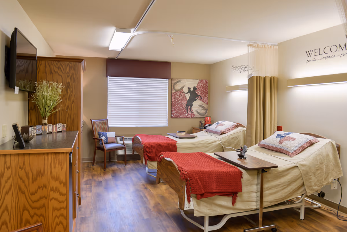 A shared bedroom in a senior living facility with two hospital-style beds, each with beige bedding and red blankets. There is a wooden cabinet with a TV mounted above it on the left side, a chair near a window with closed blinds, and a curtain divider between the beds. The walls have welcoming and home-themed decorations.