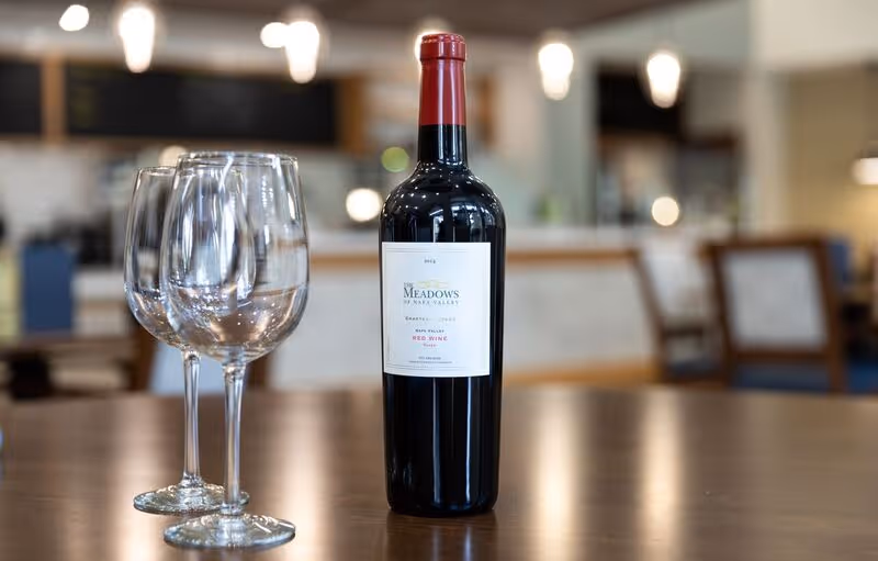 A bottle of The Meadows of Napa Valley red wine with two empty wine glasses on a wooden table in a softly lit dining area with blurred background.