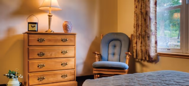 A cozy bedroom corner featuring a wooden chest of drawers with a lamp, a decorative vase, and a small framed picture on top. Next to the chest is a cushioned wooden armchair with blue upholstery. A window with floral curtains lets in natural light, and part of a bed with a quilted cover is visible in the foreground.