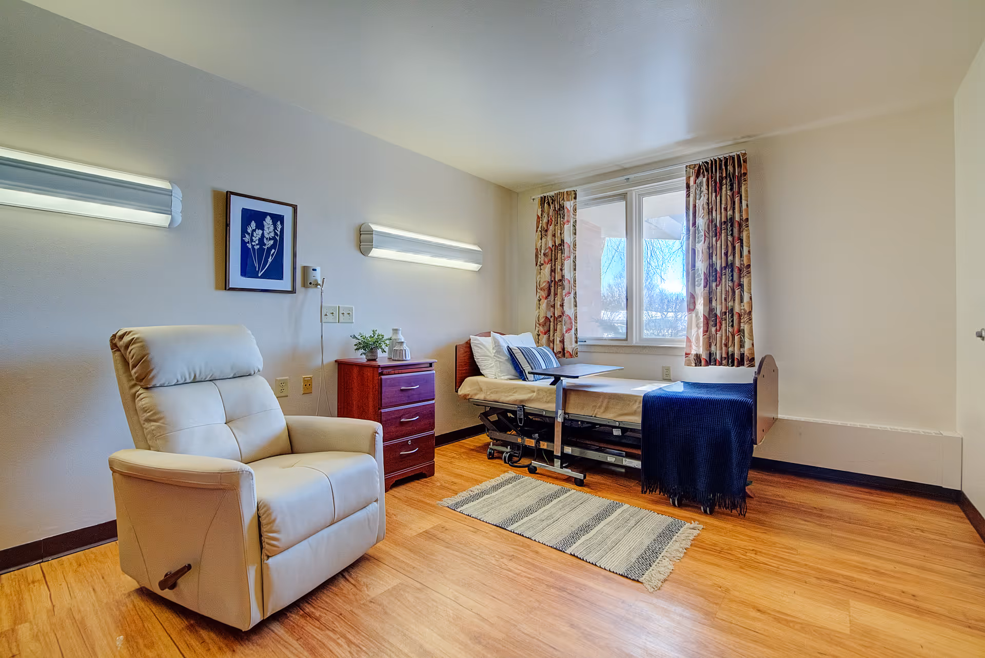 A bright and clean nursing facility room with a hospital-style bed next to a window with floral curtains. There is a beige recliner chair, a small wooden dresser with a plant and a decorative item, two wall-mounted lights, and a striped rug on the wooden floor.