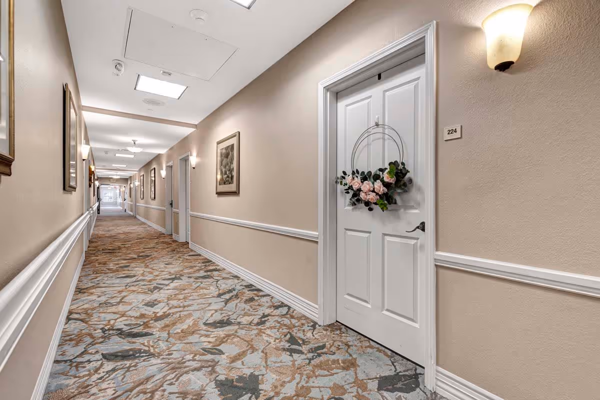 A long, well-lit hallway in a senior living facility with beige walls, white trim, and patterned carpet. Several white doors line the hallway, one of which is decorated with a circular floral wreath. Wall sconces and framed artwork are visible along the walls.