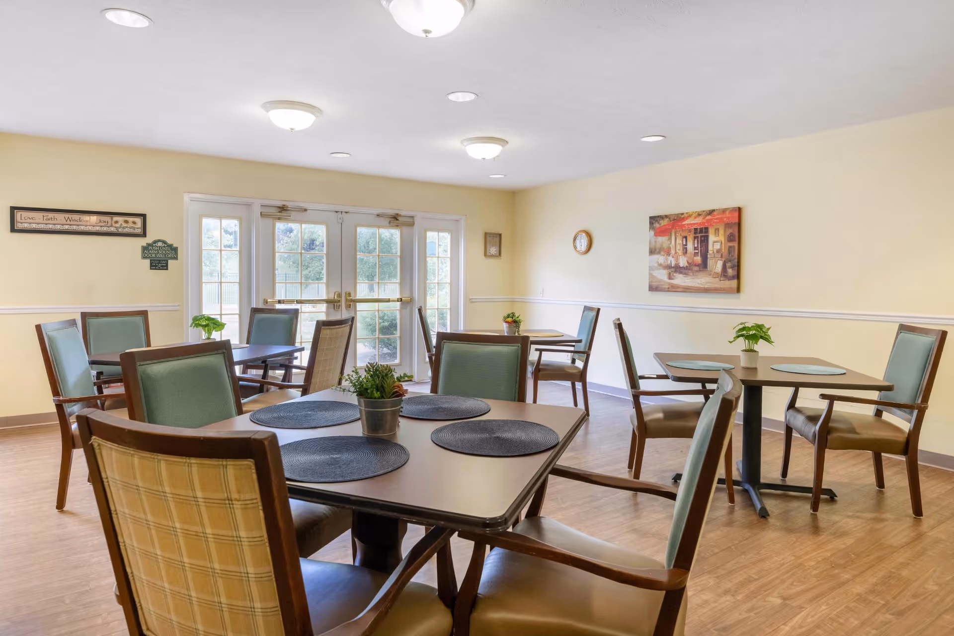 A bright dining room with several square tables, each surrounded by four chairs with green upholstery. Each table has placemats and small potted plants as centerpieces. The room has light yellow walls, wood flooring, and a set of double glass doors letting in natural light. Wall decorations include a framed picture and a small clock.