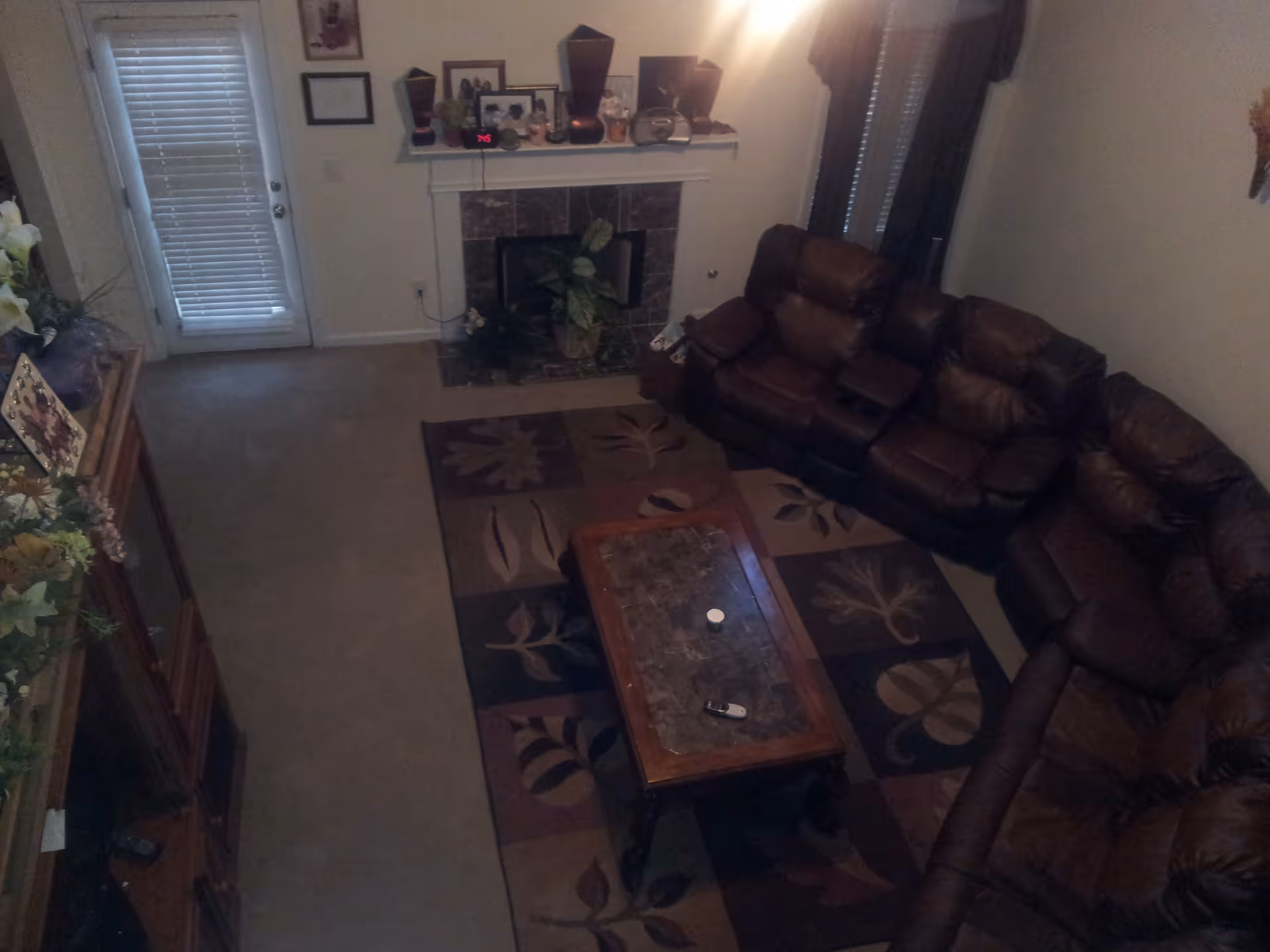 A living room with a large brown leather sectional sofa arranged around a rectangular coffee table with a marble top. The floor has a patterned area rug with leaf designs. There is a fireplace with a mantel decorated with various items, including framed photos and vases. A door with blinds is visible on the left side, and a window with dark curtains is on the right side.